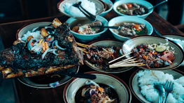 A variety of dishes are arranged on a wooden table, featuring grilled meat skewers, a large piece of roasted ribs garnished with onions and peppers, bowls of soup, and plates of white rice. The presentation is rustic with a focus on hearty, flavorful food selections.