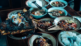A variety of dishes are arranged on a wooden table, featuring grilled meat skewers, a large piece of roasted ribs garnished with onions and peppers, bowls of soup, and plates of white rice. The presentation is rustic with a focus on hearty, flavorful food selections.