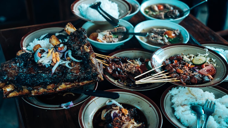A variety of dishes are arranged on a wooden table, featuring grilled meat skewers, a large piece of roasted ribs garnished with onions and peppers, bowls of soup, and plates of white rice. The presentation is rustic with a focus on hearty, flavorful food selections.