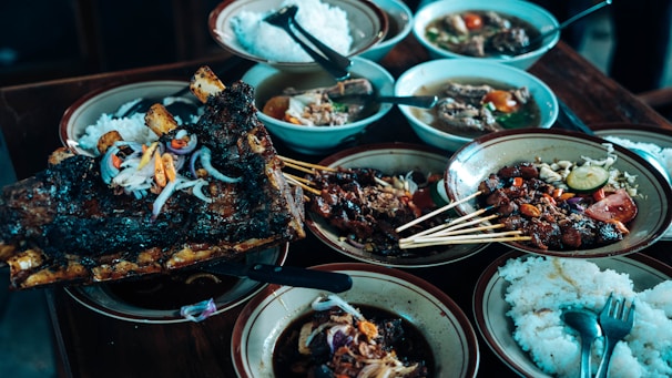 A variety of dishes are arranged on a wooden table, featuring grilled meat skewers, a large piece of roasted ribs garnished with onions and peppers, bowls of soup, and plates of white rice. The presentation is rustic with a focus on hearty, flavorful food selections.