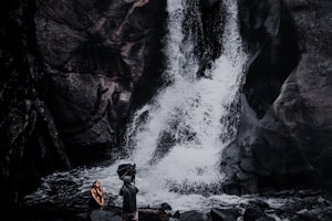 A photographer captures a person sitting near a dramatic waterfall. The rocky terrain and cascading water form a striking backdrop, while the person appears contemplative. The scene is rich with natural elements, giving an adventurous and serene feel.