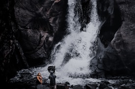 A photographer captures a person sitting near a dramatic waterfall. The rocky terrain and cascading water form a striking backdrop, while the person appears contemplative. The scene is rich with natural elements, giving an adventurous and serene feel.