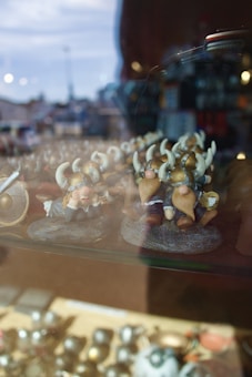 A collection of small figurines depicting Viking characters with helmets and horns is displayed on a glass shelf. The background is blurred, showing hints of an outdoor scene with blue skies and some urban elements.