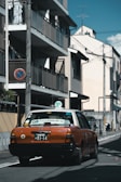 A taxi with an orange and white color scheme is driving down a narrow urban street. The street is lined with buildings, utility poles, and wires overhead. There is a no parking sign on a utility pole.