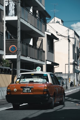 A taxi with an orange and white color scheme is driving down a narrow urban street. The street is lined with buildings, utility poles, and wires overhead. There is a no parking sign on a utility pole.