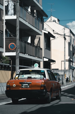 A taxi with an orange and white color scheme is driving down a narrow urban street. The street is lined with buildings, utility poles, and wires overhead. There is a no parking sign on a utility pole.