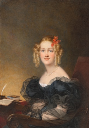 A young woman with blonde curly hair adorned with red flowers sits in an elegant chair. She wears a dark, sheer dress with puffed sleeves and a simple gold necklace. Beside her is a table with an open book, a closed book, and a quill pen in an inkstand. The background is a soft blur of neutral tones.
