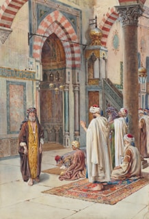 An interior scene of a religious or cultural gathering taking place in an ornately decorated building. Several men are dressed in traditional attire, some standing while others sit on colorful rugs. The architecture features arched doorways, intricate patterns, and decorative columns, indicating an Islamic style. The overall atmosphere suggests a place of worship or significant cultural importance.