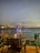 A serene waterfront view featuring a small round table with a reserved sign, candle, and chairs. The background displays a beautifully illuminated bridge with reflections on the water, set against the backdrop of a cityscape under a twilight sky.