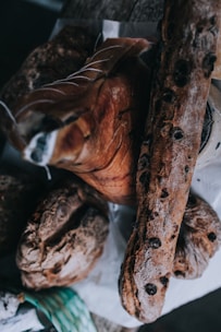An assortment of artisanal bread and cured meats is arranged on a rustic wooden surface. The bread loaves are dark and crusty, with one loaf featuring visible olives. They are accompanied by a large piece of cured meat, displaying fine marbling and intricate cutting lines.