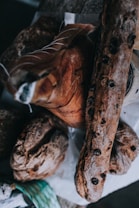 An assortment of artisanal bread and cured meats is arranged on a rustic wooden surface. The bread loaves are dark and crusty, with one loaf featuring visible olives. They are accompanied by a large piece of cured meat, displaying fine marbling and intricate cutting lines.