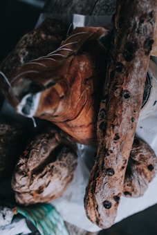 An assortment of artisanal bread and cured meats is arranged on a rustic wooden surface. The bread loaves are dark and crusty, with one loaf featuring visible olives. They are accompanied by a large piece of cured meat, displaying fine marbling and intricate cutting lines.