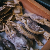 A close-up of neatly arranged uncooked pasta on a surface. The pasta appears homemade, with a dusting of flour and a rustic, artisanal appearance. The pasta shapes include folded and twisted forms with visible specks in the dough.