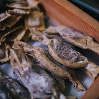 A close-up of neatly arranged uncooked pasta on a surface. The pasta appears homemade, with a dusting of flour and a rustic, artisanal appearance. The pasta shapes include folded and twisted forms with visible specks in the dough.