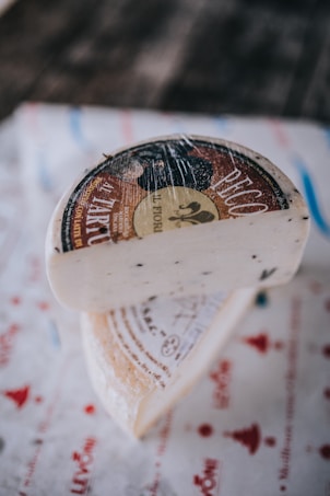 A wedge of cheese with a rustic, textured rind rests on a piece of printed paper. The cheese has a creamy white interior with fine specks, and the rind features an ornate label with text and images.