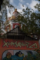 A vibrant church dome with golden accents and intricate detailing is partially obscured by lush green trees. Below, a colorful graffiti artwork featuring stylized figures and text is visible on a wall.