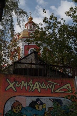 A vibrant church dome with golden accents and intricate detailing is partially obscured by lush green trees. Below, a colorful graffiti artwork featuring stylized figures and text is visible on a wall.