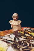 A vintage doll with a ceramic face and lace collar stands amidst a collection of colorful, detailed butterfly specimens. The butterflies feature a variety of patterns and colors, predominantly oranges, yellows, and browns. The scene is set against a dark background, creating a high contrast with the vivid colors of the butterflies and the light tones of the doll.