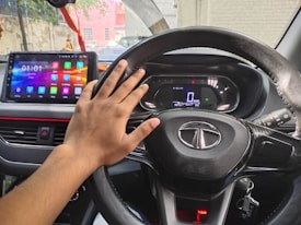 A hand is resting on the steering wheel of a car, featuring a logo. The dashboard includes a touchscreen display showing various colorful apps, and the speedometer is visible with a reading of 0 km/h. A key is inserted in the ignition, and the background outside the car shows a blurred cityscape.