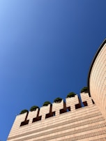 A brick structure with a modern architectural design features distinctive geometric patterns with small planted trees on top. The wall has a rounded edge that curves into the scene, and the sky is clear and blue above.