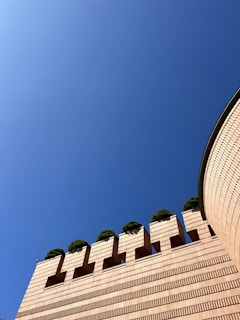 A brick structure with a modern architectural design features distinctive geometric patterns with small planted trees on top. The wall has a rounded edge that curves into the scene, and the sky is clear and blue above.