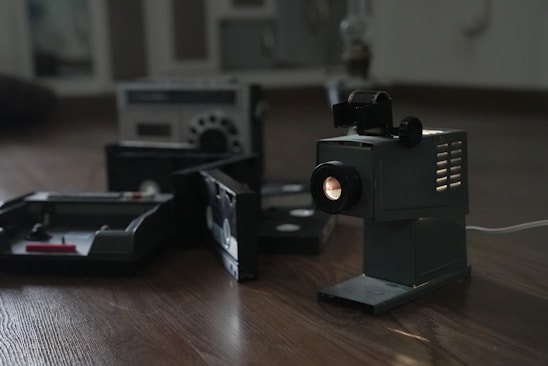A vintage film projector is positioned on a wooden floor, emitting light from its lens. In the background, there are old cassette tapes and a vintage radio or cassette player, contributing to a nostalgic atmosphere.