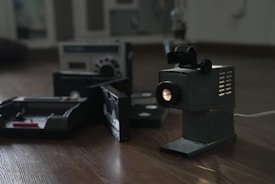 A vintage film projector is positioned on a wooden floor, emitting light from its lens. In the background, there are old cassette tapes and a vintage radio or cassette player, contributing to a nostalgic atmosphere.