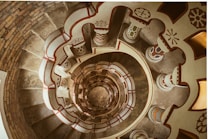 A spiral staircase viewed from above with intricately decorated columns featuring various geometric patterns. The stairs appear to be made of stone or concrete, and the embellishments show a symmetrical design. Warm lighting creates an inviting ambiance, and the repetitive spiral creates a visually pleasing depth.