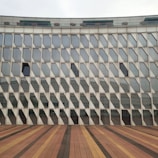 A modern architectural building facade featuring numerous geometric, diamond-shaped glass panels that create a repetitive pattern. The structure's base is not visible, but a wooden deck, composed of different shades of brown, stretches out in front.