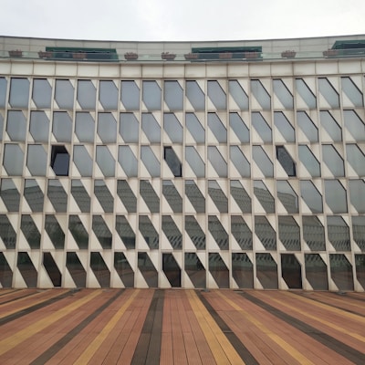 A modern architectural building facade featuring numerous geometric, diamond-shaped glass panels that create a repetitive pattern. The structure's base is not visible, but a wooden deck, composed of different shades of brown, stretches out in front.