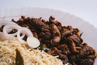 A plate of spicy, well-seasoned meat served alongside a portion of rice, garnished with sliced raw onions. The meat appears to be richly coated in dark-colored spices, and the rice is light and fluffy.