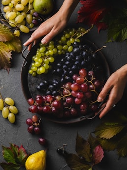 A hand is arranging a variety of grapes on a round, dark platter. The grapes are in shades of green, red, and dark purple, creating a rich color contrast. Surrounding the platter, there are additional grapes, a pear, and vibrant autumn leaves scattered on a dark textured surface.