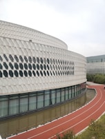 A modern architectural structure featuring a curved facade with a diamond pattern. There are large glass windows along the lower part of the building. A red running track with white markings curves in front of the building, bordered by a narrow water channel.