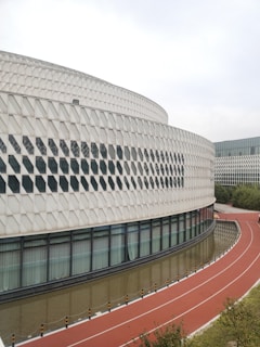 A modern architectural structure featuring a curved facade with a diamond pattern. There are large glass windows along the lower part of the building. A red running track with white markings curves in front of the building, bordered by a narrow water channel.
