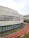 A modern architectural structure featuring a curved facade with a diamond pattern. There are large glass windows along the lower part of the building. A red running track with white markings curves in front of the building, bordered by a narrow water channel.