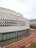A modern architectural structure featuring a curved facade with a diamond pattern. There are large glass windows along the lower part of the building. A red running track with white markings curves in front of the building, bordered by a narrow water channel.