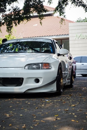 A white sports car is parked under the shade of a tree with green leaves draping over parts of the car. The background includes a tiled roof building and a glimpse of another car. The car appears sleek and polished, with black rims and a customized front bumper.
