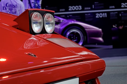A close-up of the front part of a vintage red sports car, featuring the pop-up headlights and sleek angular design. In the background, a timeline with various years is visible alongside a purple sports car.
