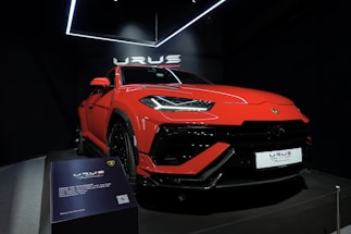 A vibrant red luxury SUV is displayed under bright showroom lights, positioned on a platform with sleek, aerodynamic lines and aggressive front profile. The branding is prominently visible above the car, and there is a plaque providing specific details about the vehicle model.