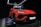 A vibrant red luxury SUV is displayed under bright showroom lights, positioned on a platform with sleek, aerodynamic lines and aggressive front profile. The branding is prominently visible above the car, and there is a plaque providing specific details about the vehicle model.