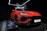 A vibrant red luxury SUV is displayed under bright showroom lights, positioned on a platform with sleek, aerodynamic lines and aggressive front profile. The branding is prominently visible above the car, and there is a plaque providing specific details about the vehicle model.