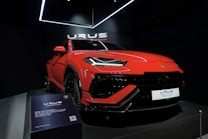 A vibrant red luxury SUV is displayed under bright showroom lights, positioned on a platform with sleek, aerodynamic lines and aggressive front profile. The branding is prominently visible above the car, and there is a plaque providing specific details about the vehicle model.