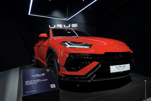 A vibrant red luxury SUV is displayed under bright showroom lights, positioned on a platform with sleek, aerodynamic lines and aggressive front profile. The branding is prominently visible above the car, and there is a plaque providing specific details about the vehicle model.