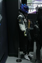 A mannequin in a racing suit and blue helmet is leaning against a black wall. Various racing logos are displayed on the suit, and there are trophies in the background. A person with a backpack is standing nearby.