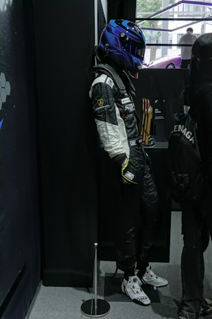 A mannequin in a racing suit and blue helmet is leaning against a black wall. Various racing logos are displayed on the suit, and there are trophies in the background. A person with a backpack is standing nearby.