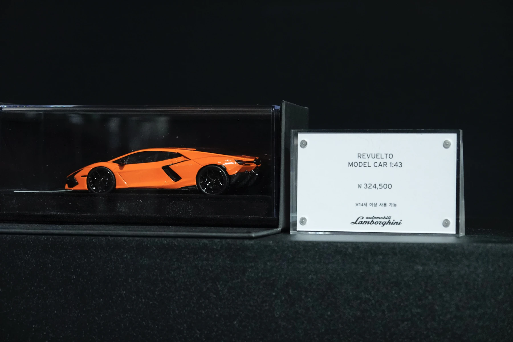 An orange model sports car in a clear display case beside its name card, on a dark shelf