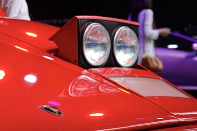 A close-up of a vintage car's headlight assembly, featuring two circular headlights embedded in a rectangular enclosure on a sleek red car body. The prominent logo displayed on the car indicates its luxury sports car brand. The background includes an out-of-focus person and a purple vehicle, suggesting a showroom or exhibit environment.