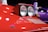 A close-up of a vintage car's headlight assembly, featuring two circular headlights embedded in a rectangular enclosure on a sleek red car body. The prominent logo displayed on the car indicates its luxury sports car brand. The background includes an out-of-focus person and a purple vehicle, suggesting a showroom or exhibit environment.