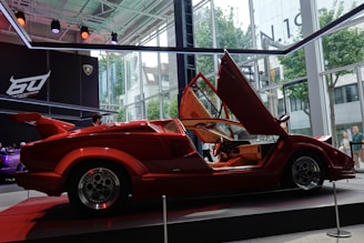 A sleek red sports car with distinctive scissor doors open, showcased in a modern showroom. The car is set against a backdrop of glass walls showing urban architecture and trees, with additional lighting focused on the vehicle. Branding elements and logos are visible in the background.