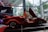 A sleek red sports car with distinctive scissor doors open, showcased in a modern showroom. The car is set against a backdrop of glass walls showing urban architecture and trees, with additional lighting focused on the vehicle. Branding elements and logos are visible in the background.